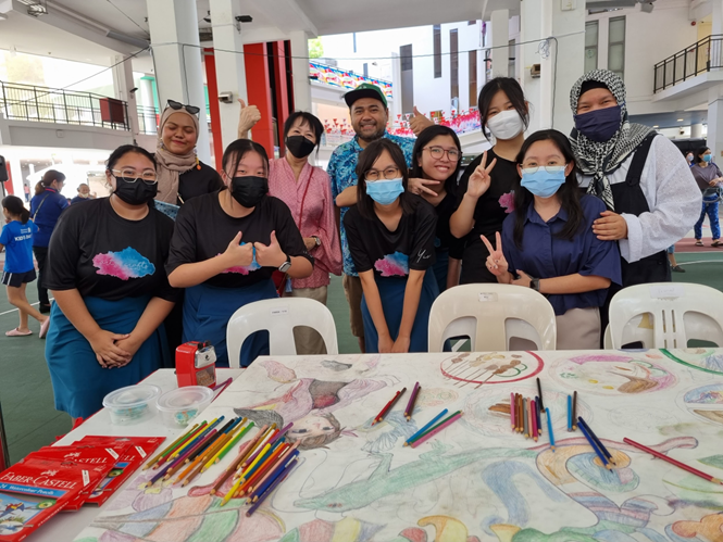 Group photo of students and adults gathered around a table covered with colorful drawings and pencils during an art activity session